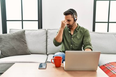 Young hispanic man with beard wearing call center agent headset working from home tired rubbing nose and eyes feeling fatigue and headache. stress and frustration concept. 