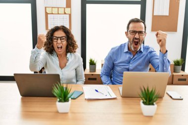 Middle age hispanic woman and man sitting with laptop at the office angry and mad raising fist frustrated and furious while shouting with anger. rage and aggressive concept. 