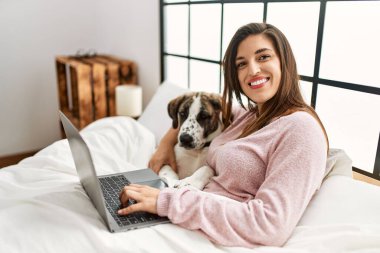 Yatak odasında dizüstü bilgisayar kullanan, köpeğini kucaklayan genç bir kadın.