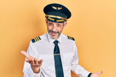 Yakışıklı, orta yaşlı, gri saçlı, uçak pilotu üniforması giyen, neşeli, kollarını açıp gülümseyen, dost canlısı, olumlu ve kendinden emin bir şekilde selamlayan bir adam. 