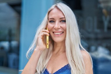 Genç sarışın kadın gülümsüyor. Kendine güveni tam. Akıllı telefondan konuşuyor.