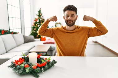 Arap genç adam Noel ağacının yanında oturmuş gururla gülümseyen kol kaslarını gösteriyor. fitness konsepti. 