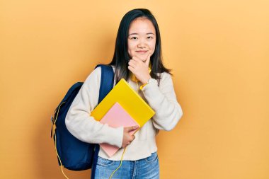 Young chinese girl holding student backpack and books looking confident at the camera smiling with crossed arms and hand raised on chin. thinking positive. 