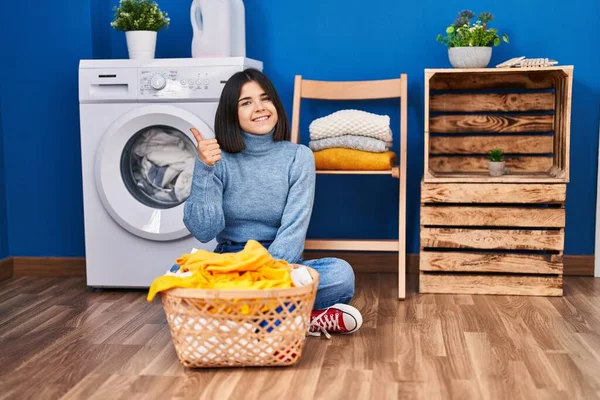 Young hispanic woman at laundry room smiling happy and positive, thumb up doing excellent and approval sign