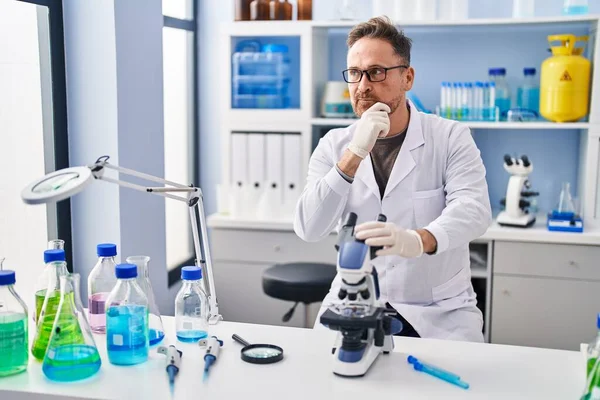 Middle age caucasian man working at scientist laboratory serious face thinking about question with hand on chin, thoughtful about confusing idea