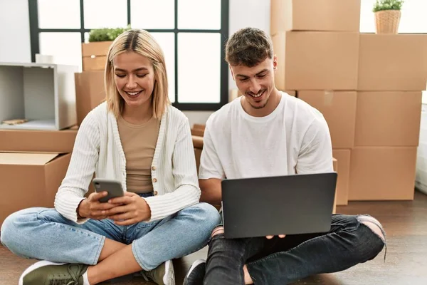 一对年轻的高加索夫妇坐在新家 一边笑着一边用笔记本电脑和智能手机 — 图库照片