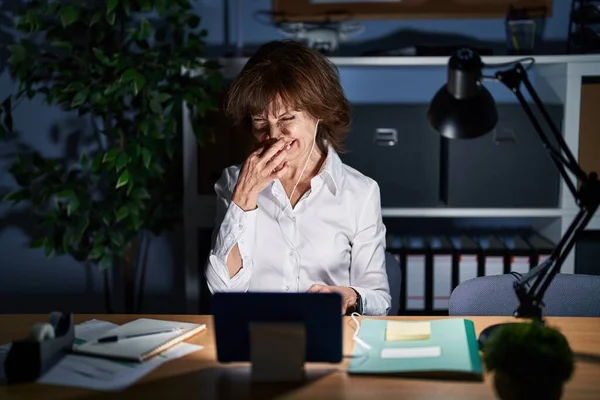 Middle age woman working at the office at night smelling something stinky and disgusting, intolerable smell, holding breath with fingers on nose. bad smell