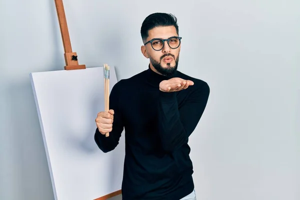 Handsome man with beard holding brushes close to easel stand looking at the camera blowing a kiss with hand on air being lovely and sexy. love expression.