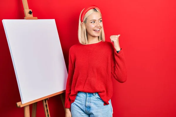 Beautiful Blonde Woman Standing Painter Easel Stand Pointing Thumb Side — Foto de Stock