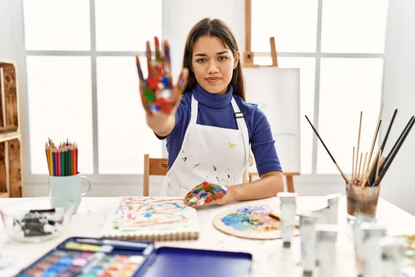 Young brunette woman at art studio with painted hands doing stop sing with palm of the hand. warning expression with negative and serious gesture on the face.