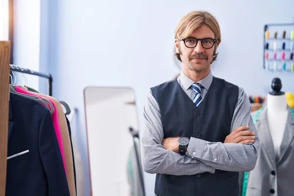 Young blond man tailor smiling confident standing with arms crossed gesture at clothing factory