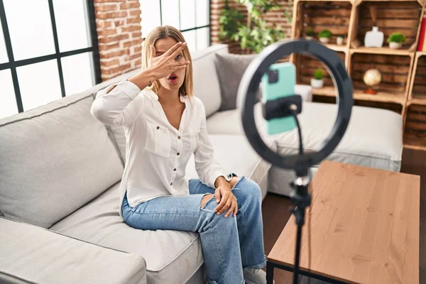 Young blonde woman recording vlog tutorial with smartphone at home peeking in shock covering face and eyes with hand, looking through fingers afraid 