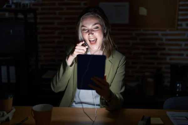 Blonde caucasian woman working at the office at night pointing to you and the camera with fingers, smiling positive and cheerful