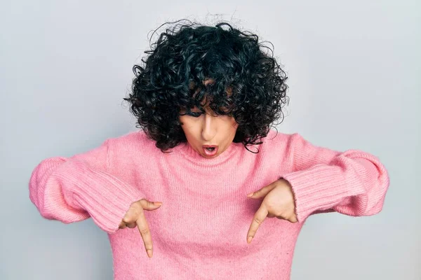 Young middle east woman wearing casual clothes pointing down with fingers showing advertisement, surprised face and open mouth 