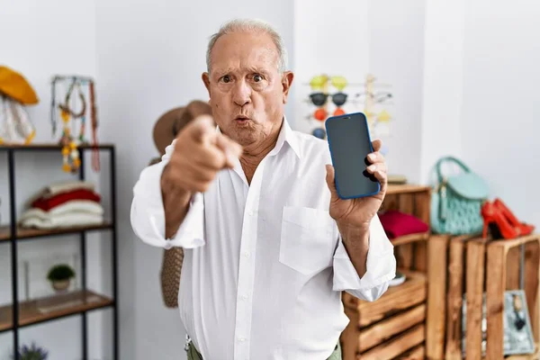 Senior man holding smartphone at retail shop pointing with finger to the camera and to you, confident gesture looking serious