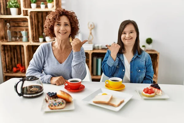 Family of mother and down syndrome daughter sitting at home eating breakfast smiling with happy face looking and pointing to the side with thumb up.