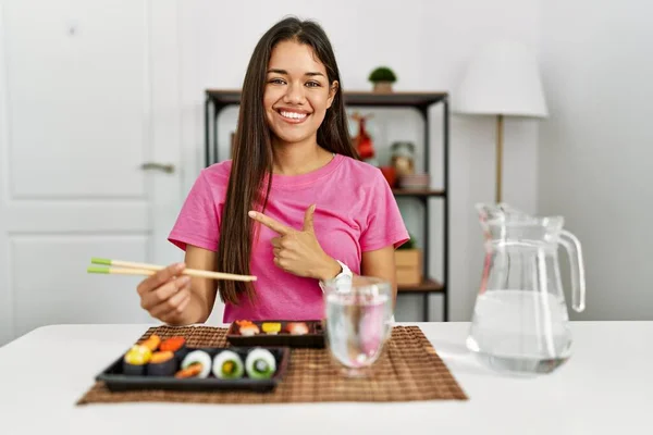 若いブルネットの女性が幸せと自然な表情で側に手と指を指して顔に笑顔で陽気な箸を使用して寿司を食べる — ストック写真