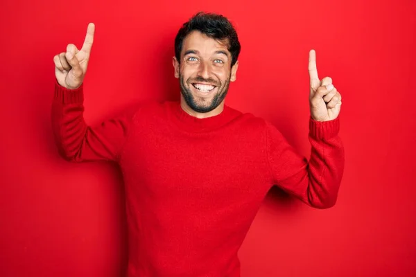 髭を生やしたハンサムな男はカジュアルな赤いセーターを身に着けている笑顔と驚きと指で指摘し 腕を上げ — ストック写真