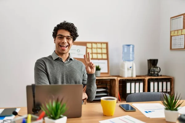 Young hispanic man wearing business style sitting on desk at office smiling with happy face winking at the camera doing victory sign. number two.
