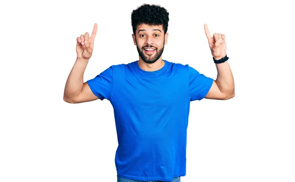 年轻的阿拉伯男子 留着胡须 穿着休闲的蓝色T恤 面带微笑 惊讶而惊讶 用手指和胳膊指指着 — 图库照片