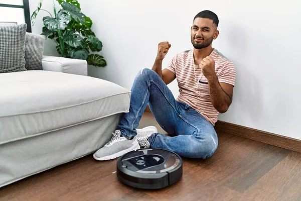 Young indian man sitting at home by vacuum robot very happy and excited doing winner gesture with arms raised, smiling and screaming for success. celebration concept.