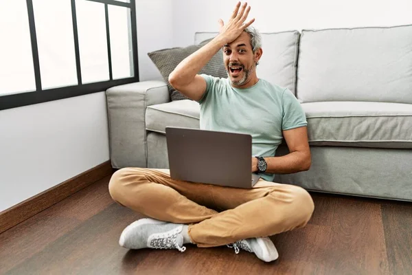 Middle age hispanic man using laptop sitting on the floor at the living room surprised with hand on head for mistake, remember error. forgot, bad memory concept.