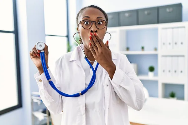 Black doctor woman holding stethoscope covering mouth with hand, shocked and afraid for mistake. surprised expression