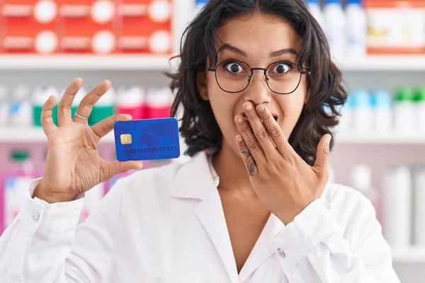 薬局で働いている若いヒスパニック系の女性は 手で口をカバークレジットカードを持っています ショックを受け 間違いを恐れて 驚きの表情 — ストック写真