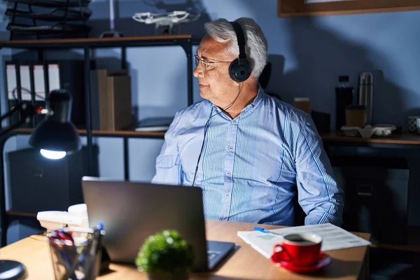 Hispanic senior man wearing call center agent headset at night looking to side, relax profile pose with natural face with confident smile.