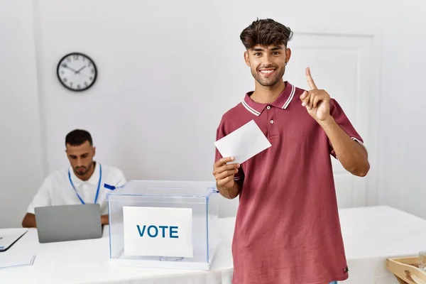 幸せな顔で指を指すアイデアや質問に驚いて投票箱に封筒を入れて若いヒスパニック系の男性投票 ナンバーワン — ストック写真