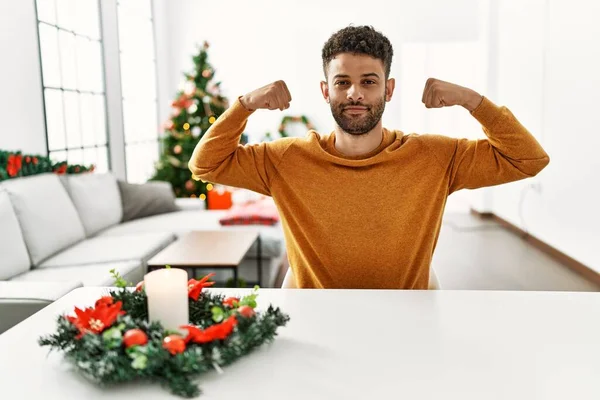 Arap genç adam Noel ağacının yanında oturmuş gururla gülümseyen kol kaslarını gösteriyor. fitness konsepti. 