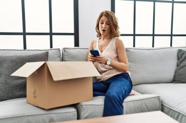 Orta yaşlı bir kadın kanepede oturmuş telefonda teslimat paketini kontrol ediyor. Korkmuş ve ağzı açık bir şekilde şaşırmış. 