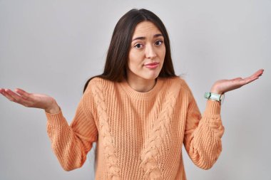 Beyaz arka planda duran esmer bir kadın. Kolları ve elleri havada, kafası karışmış bir ifade. şüphe kavramı. 