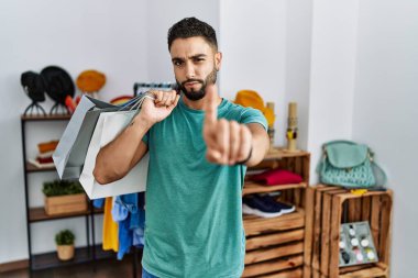 Satış mağazasında sakallı, alışveriş torbaları tutan genç ve yakışıklı bir adam parmağını kaldırmış ve kızgın bir ifadeyle işaret ediyor. 