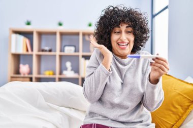 Young brunette woman with curly hair holding pregnancy test result celebrating victory with happy smile and winner expression with raised hands 