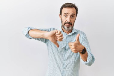 Middle age hispanic man with beard standing over isolated background doing thumbs up and down, disagreement and agreement expression. crazy conflict 