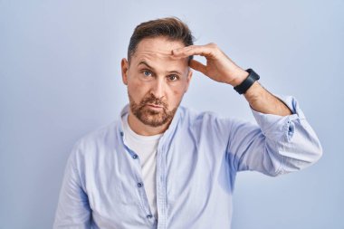 Middle age caucasian man standing over blue background worried and stressed about a problem with hand on forehead, nervous and anxious for crisis 