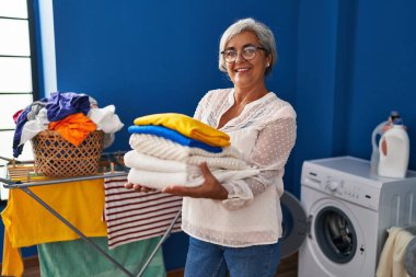 Çamaşırhanede katlanmış giysileri tutan orta yaşlı bir kadın gülümsüyor.
