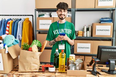 Young hispanic man wearing volunteer uniform usong smartphone at charity center.