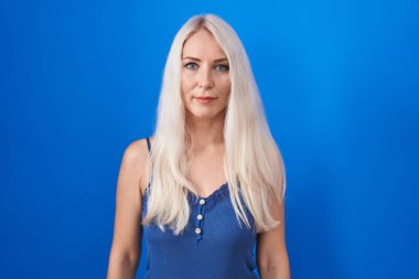 Caucasian woman standing over blue background relaxed with serious expression on face. simple and natural looking at the camera. 