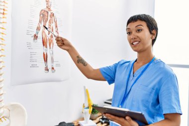 Young hispanic woman wearing physiotherapist uniform pointing to vertebral column holding checklist at physiotherapy clinic
