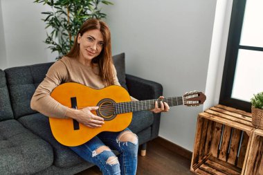 Orta yaşlı beyaz kadın klasik gitar çalıyor. Evdeki kanepede oturuyor..