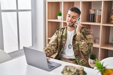 Genç İspanyol asker dizüstü bilgisayar kullanıyor. Evdeki akıllı telefondan konuşuyor.