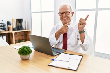 Senior man working at the office using computer laptop smiling and looking at the camera pointing with two hands and fingers to the side. 