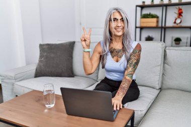 Middle age grey-haired woman using laptop at home showing and pointing up with fingers number two while smiling confident and happy. 