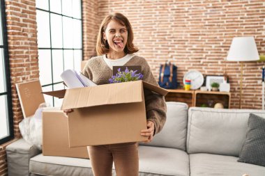 Young beautiful woman holding box moving to a new home sticking tongue out happy with funny expression. 