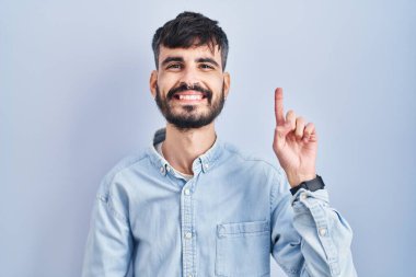 Young hispanic man with beard standing over blue background showing and pointing up with finger number one while smiling confident and happy. 