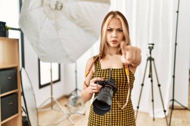 Young caucasian photographer girl holding professional camera at photography studio pointing with finger to the camera and to you, confident gesture looking serious 