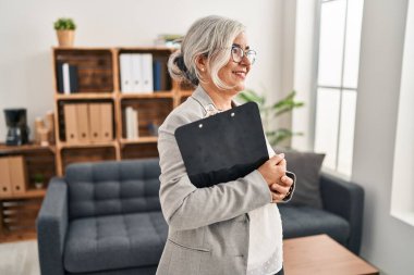 Middle age woman with grey hair at consultation office looking to side, relax profile pose with natural face and confident smile. 