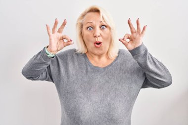 Middle age caucasian woman standing over white background looking surprised and shocked doing ok approval symbol with fingers. crazy expression 